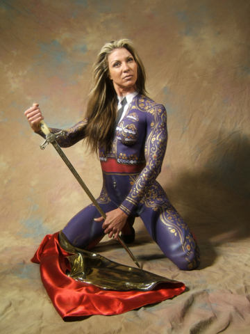 long haired woman in full body paint kneeling in a purple matador outfit holding a sword with a red flag on the floor below her